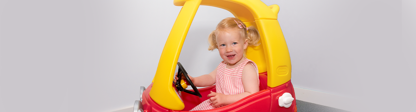 Young child sitting in a red and yellow toy car, holding the steering wheel and facing the camera.