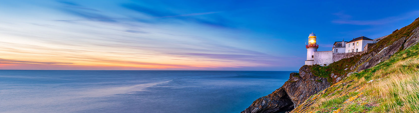 Lighthouse glowing on a rocky cliff above the ocean at sunset, with a blue‑to‑orange sky and calm water stretching to the horizon.