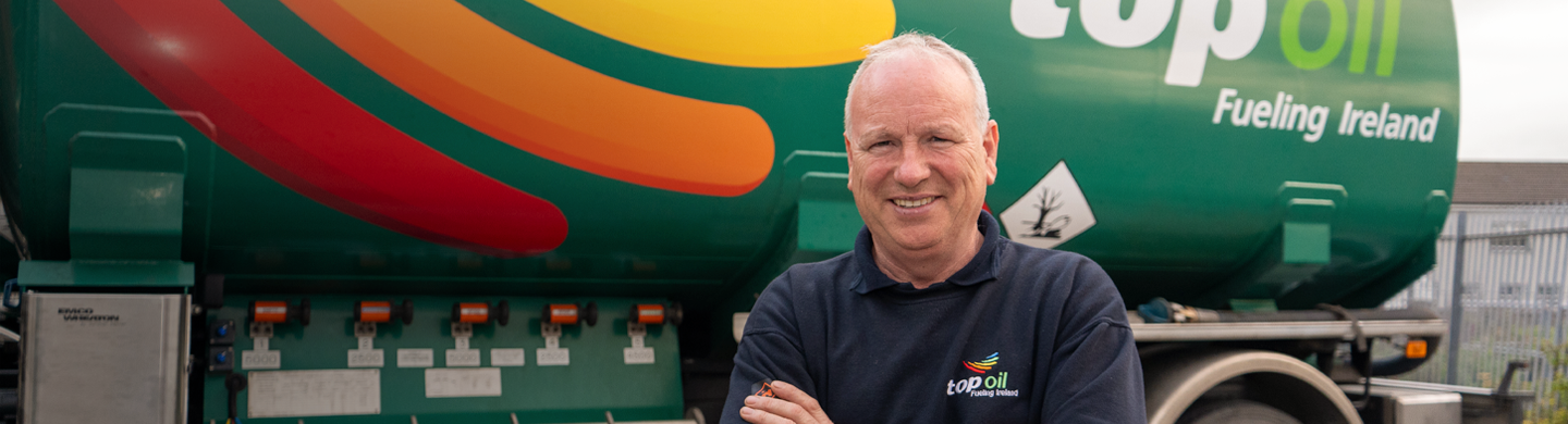 Person stands with arms crossed in front of a green Top Oil fuel truck displaying the text “Top Oil – Fueling Ireland” and multi‑color stripe graphics.