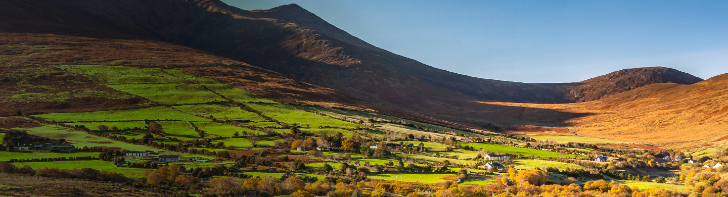 Wide landscape view of a rural valley with patchwork green fields, scattered homes, and trees beneath rolling brown mountains under a clear blue sky.
