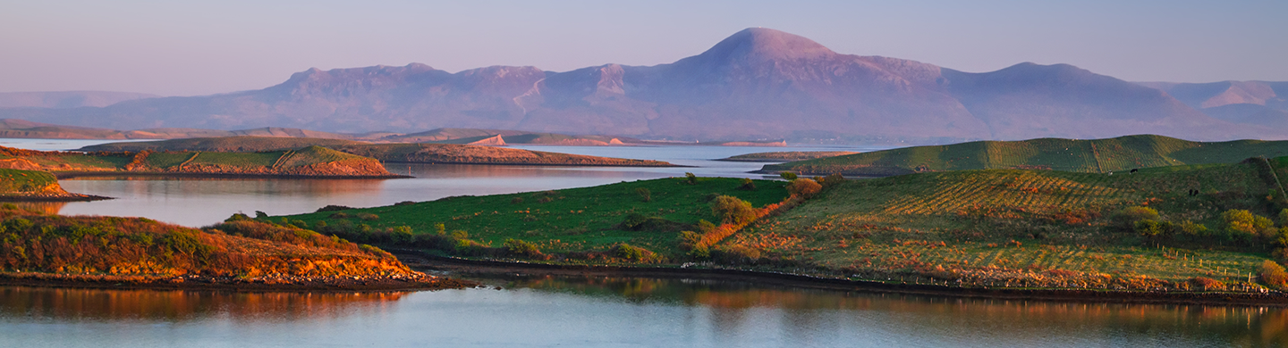 Coastal landscape with water, rolling green hills, and mountains in the background.
